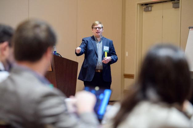 Wes Ladd stands beside a podium and speaks at a conference in a hotel ballroom.